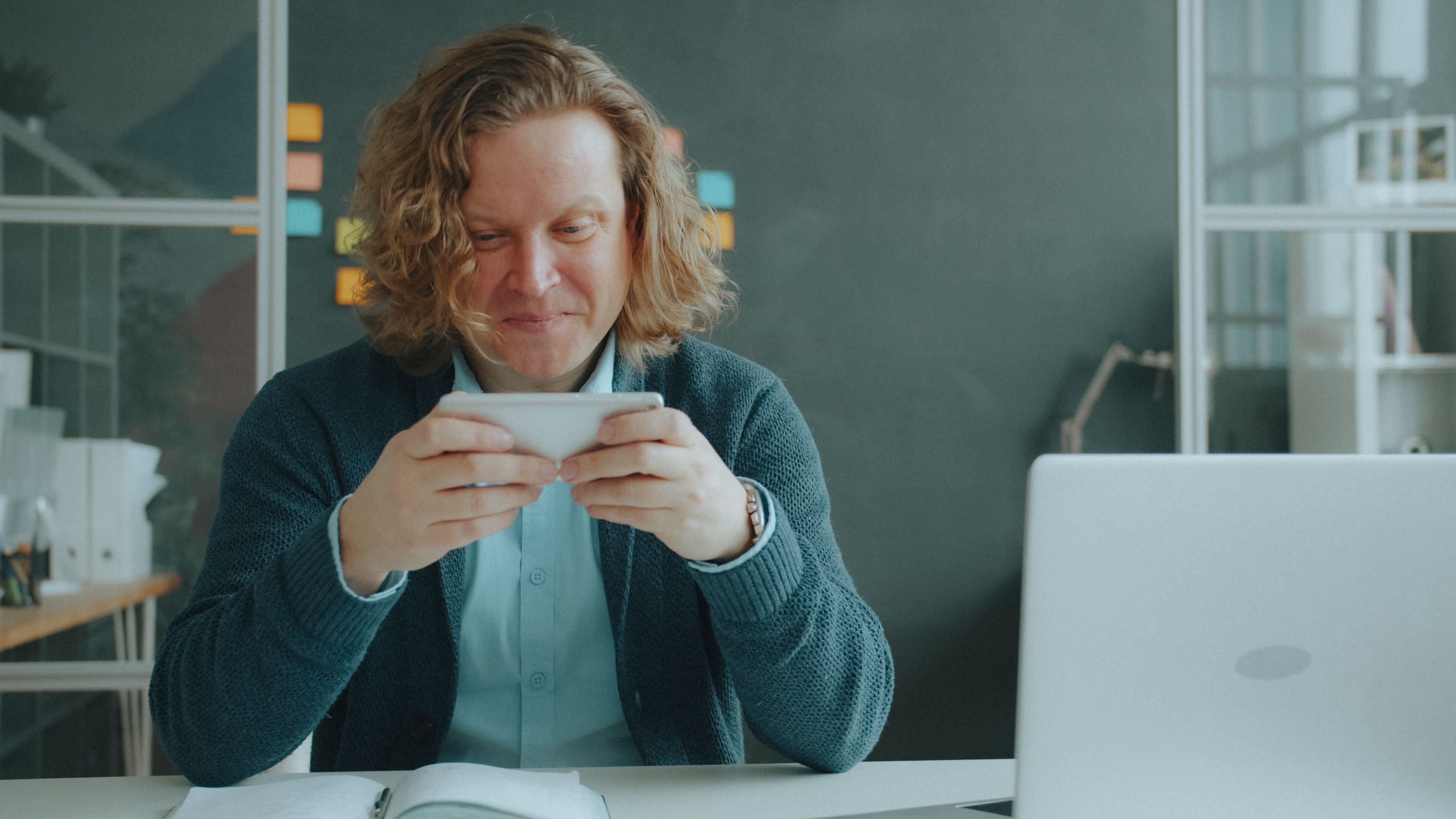 Smiling man using smartphone in a modern office environment with a laptop and open book on the desk.