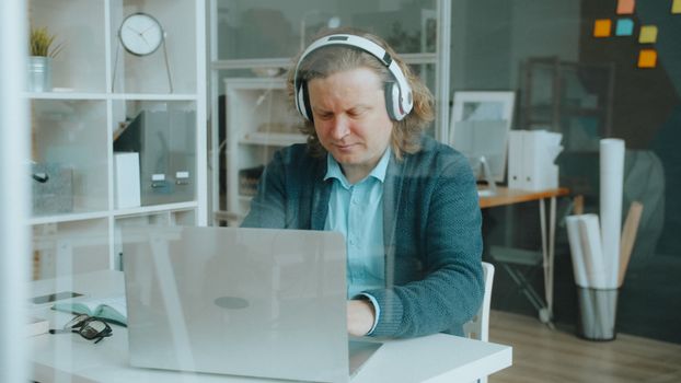 Focused man wearing headphones working on laptop in modern office setting through glass.