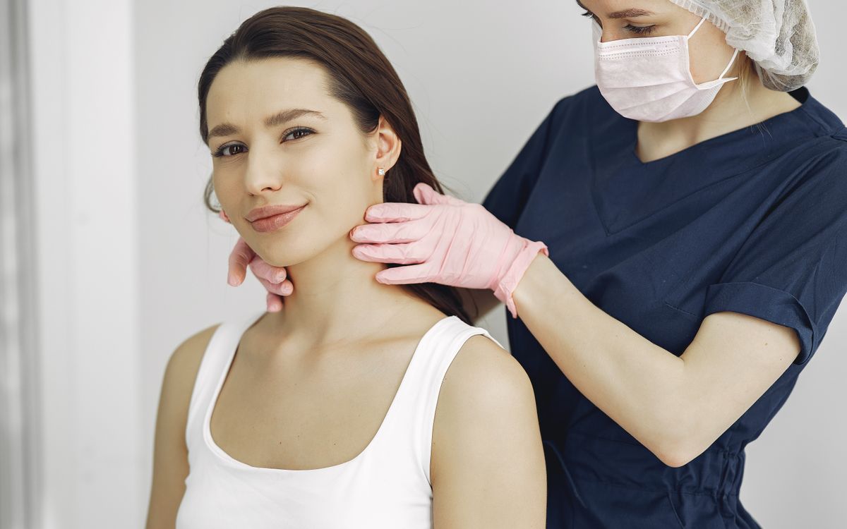 A medical practitioner in scrub suit performs a neck examination on a smiling woman in a clinic.