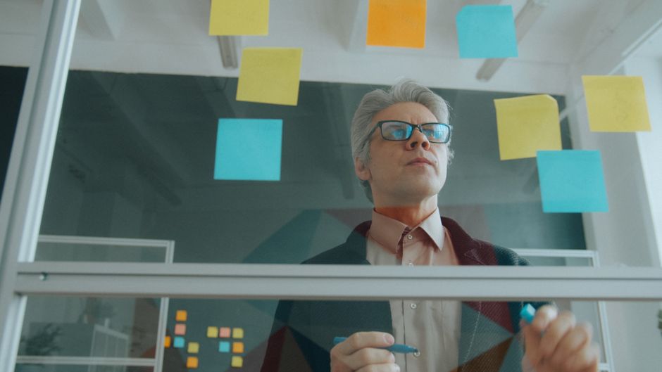Focused mature businessman strategizing with sticky notes on glass in a modern office.