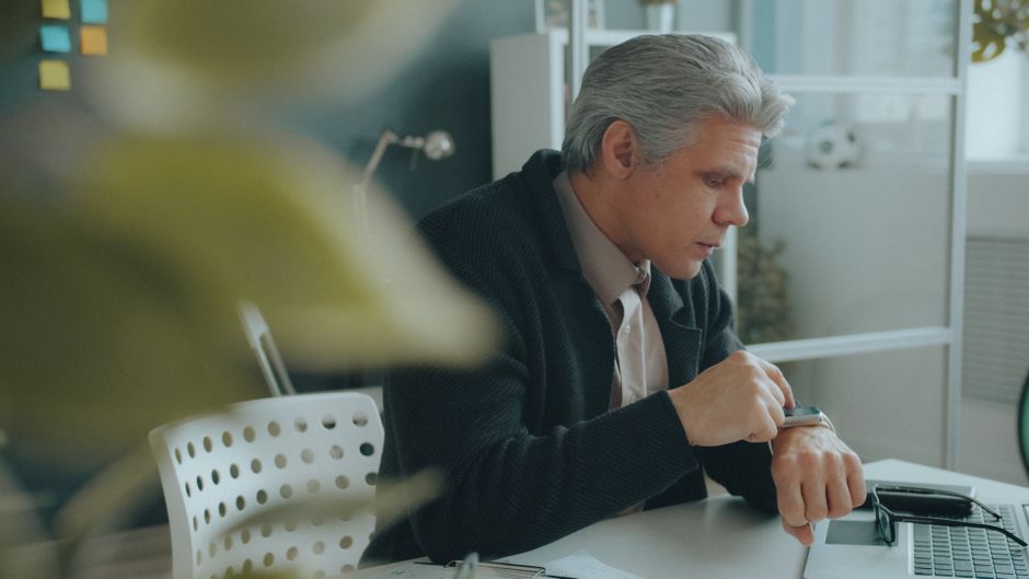Senior adult businessman looking at smartwatch while sitting at desk in modern office.