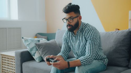 Free Bearded man with glasses enjoying video games on couch in modern living room. Stock Photo