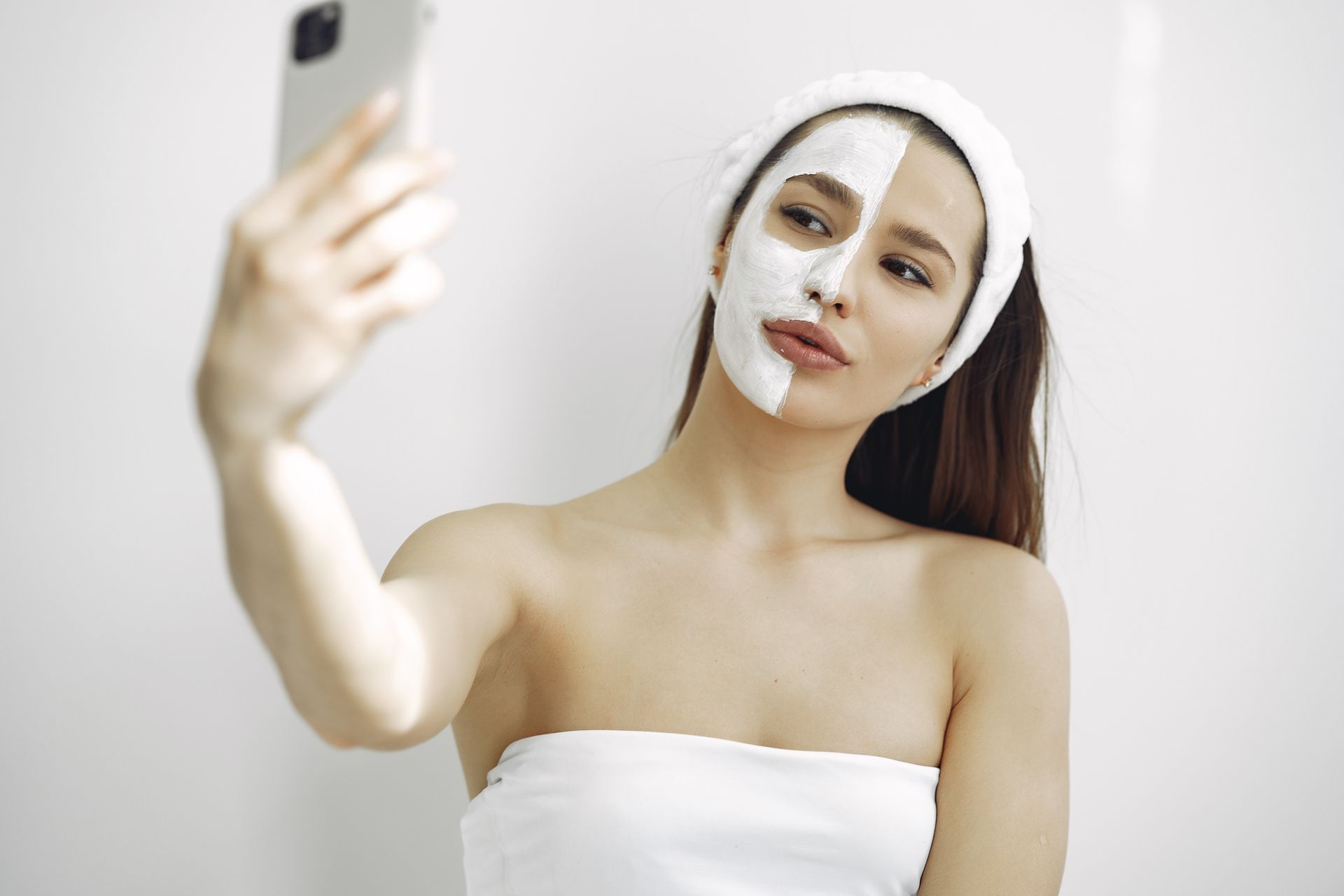 Young woman with a facial mask on half her face taking a selfie indoors. Beauty and skincare concept.