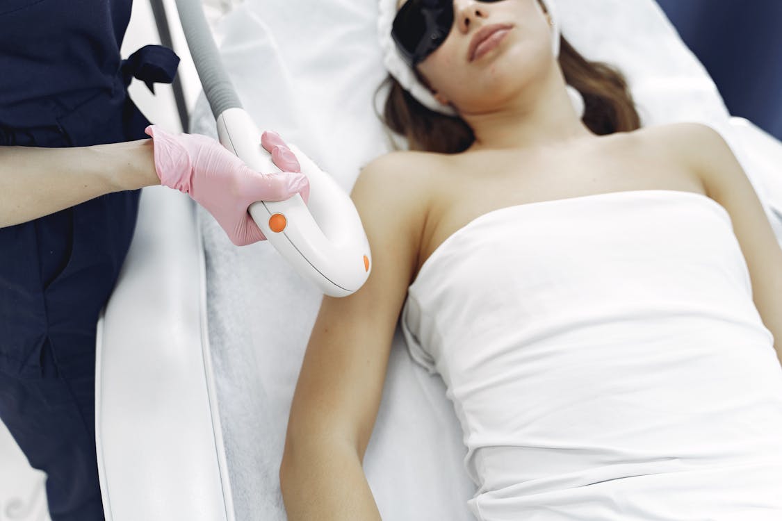 Crop faceless beautician conducting laser hair removal to young lady in safety glasses in clinic · free stock photo