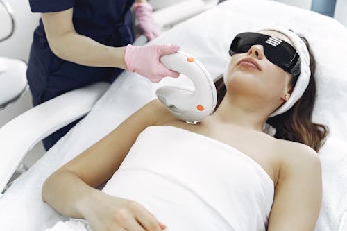 Crop faceless woman in medical uniform and gloves using laser tool on young woman wearing safety glasses and towel lying on couch in hospital