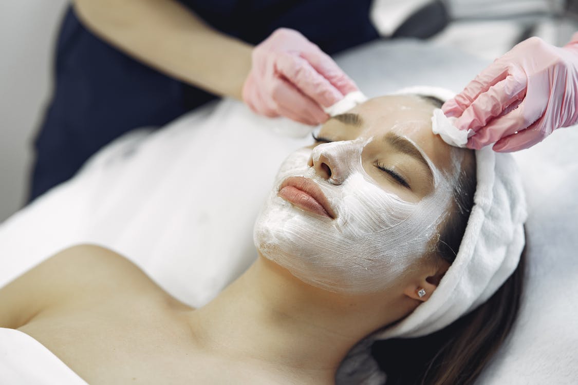 Free From above anonymous medical specialist in uniform and gloves wiping cream mask off face of relaxed woman during skincare procedure in modern studio Stock Photo