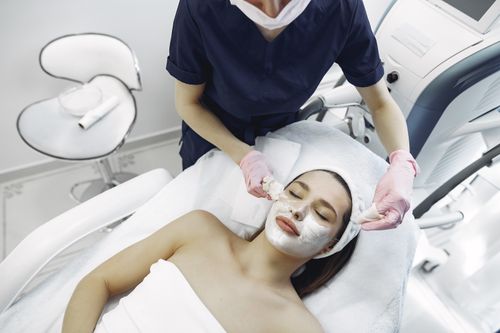 Free From above of dermatologist in medical coat and latex gloves doing face cleansing and wiping mask from face of female lying in towel in modern salon Stock Photo