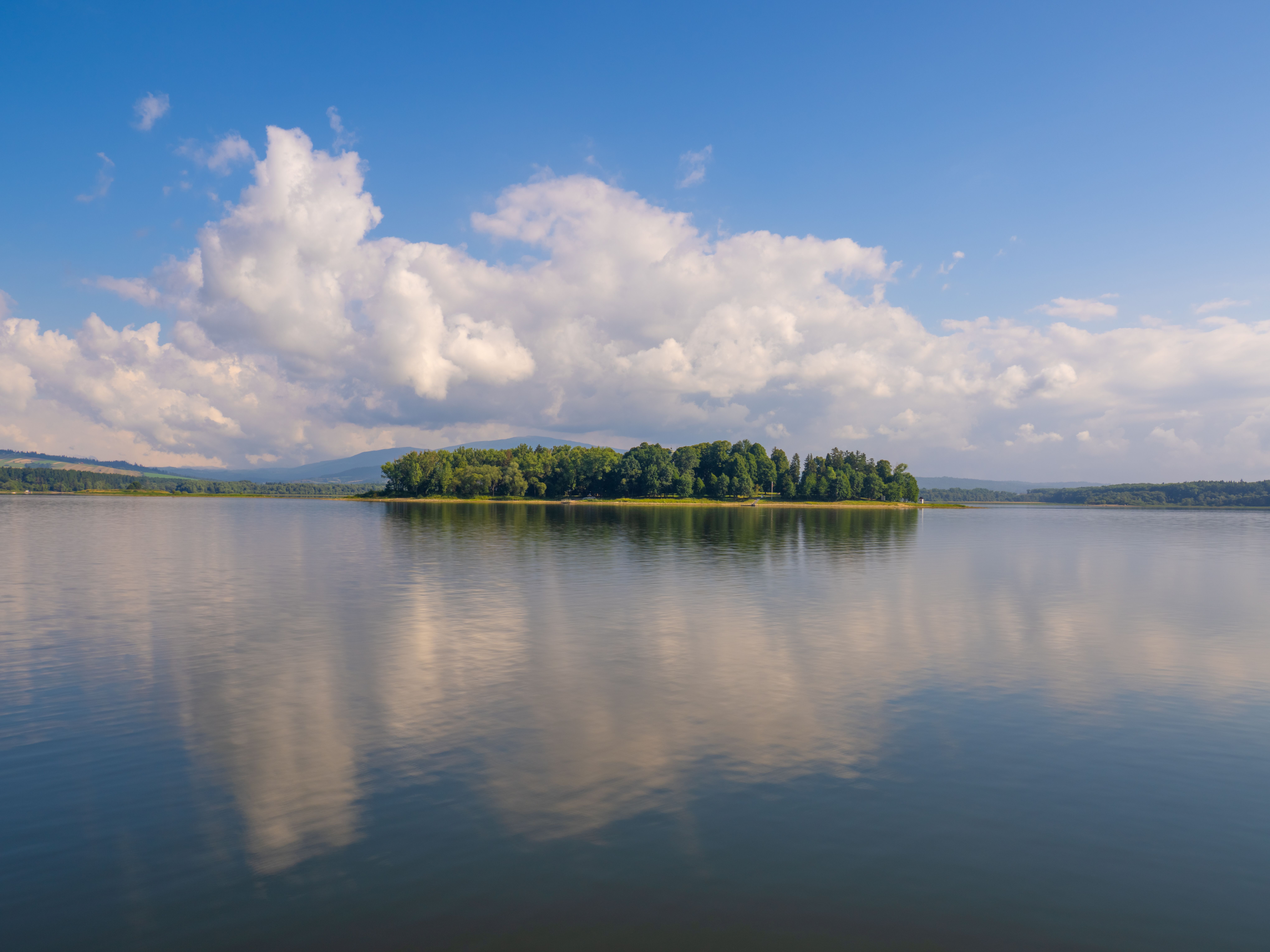 Fotomural Vista tranquila de una isla boscosa en un lago en calma con reflejos de las nubes.