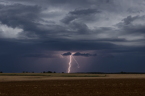 Free A powerful lightning bolt strikes during a storm, illuminating a rural landscape with dramatic clouds. Stock Photo