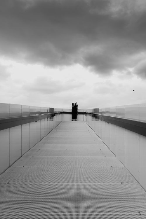 Free Black and white image of a couple embracing on a futuristic skybridge, under dramatic clouds. Stock Photo