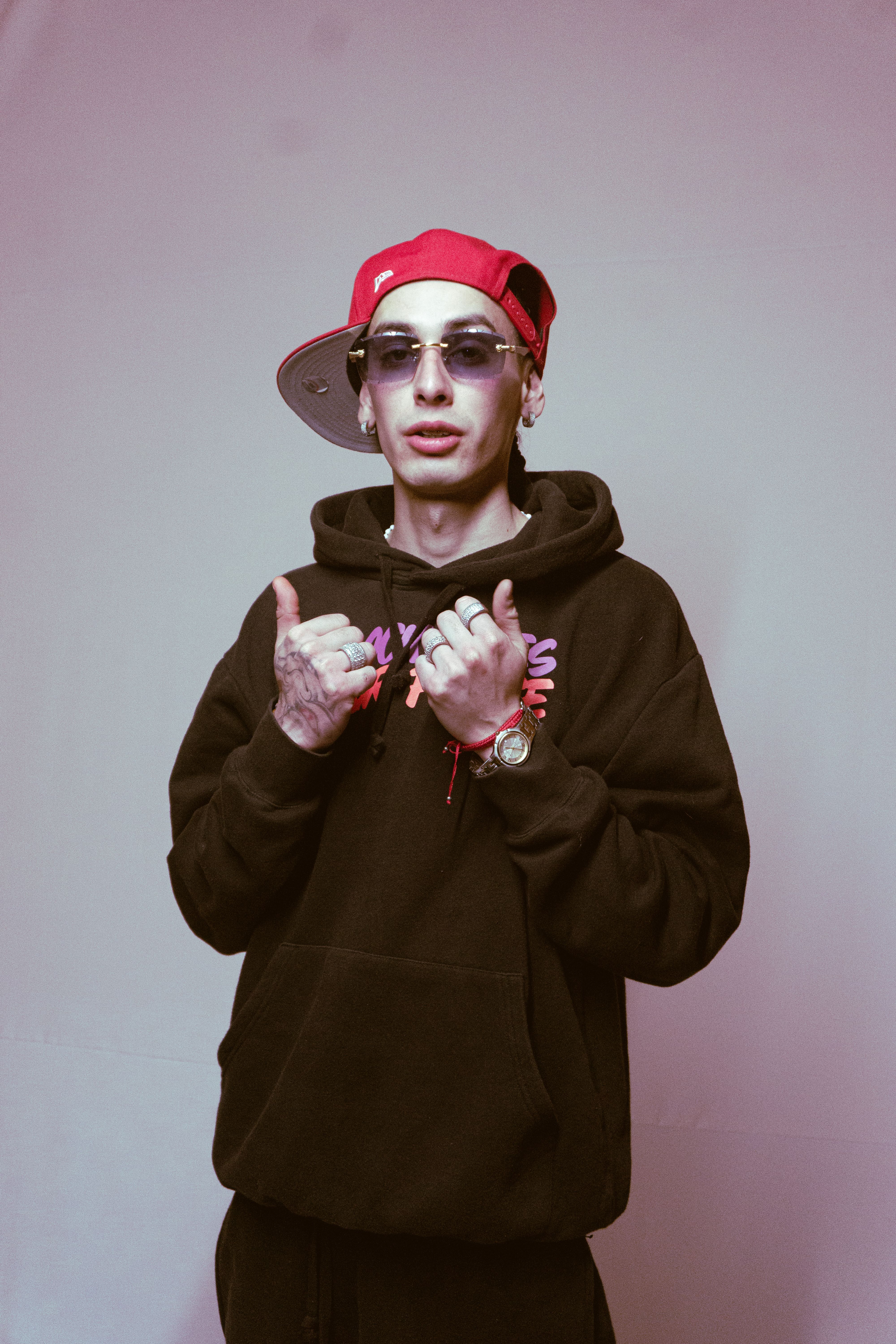 Trendy young adult wearing sunglasses and a red cap, making hand gestures. Urban fashion style.