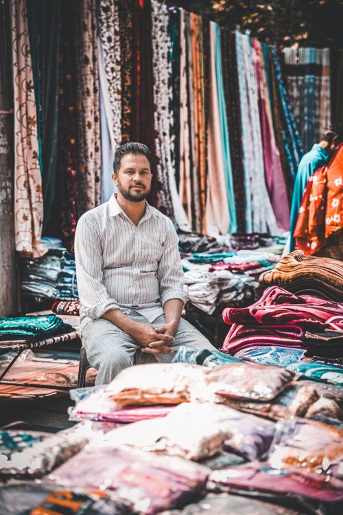 Free Man sits among colorful fabrics in an outdoor market, showcasing textiles and vibrant clothing. Stock Photo