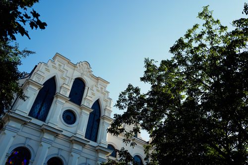 Free Elegant white church facade against blue sky, featuring arched stained glass windows and lush greenery. Stock Photo