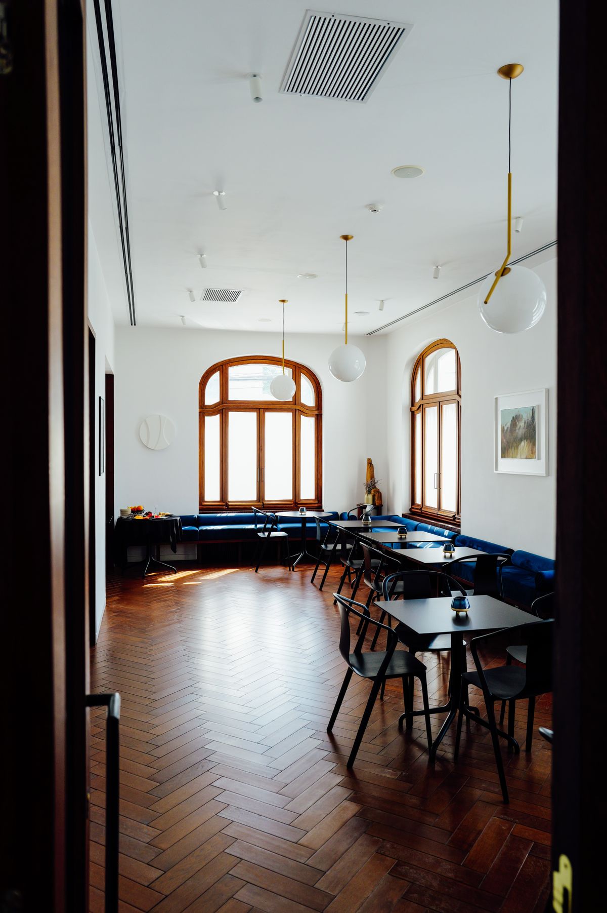 Lacivert kadife koltuklu, siyah sandalyeli masalı, kemerli pencereli ve herringbone parke zeminli bir salon; tavanda cam küre lambalar asılı.