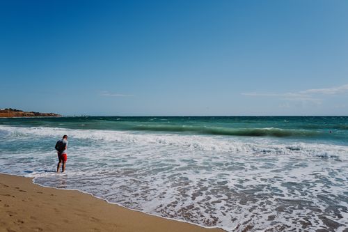 Free A man in red shorts walks along a sunny beach shoreline under a clear blue sky. Stock Photo