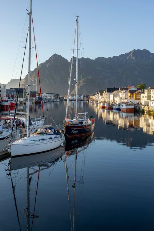 Free Scenic view of sailboats moored in Henningsvær harbor with mountains in the background and clear skies above. Stock Photo