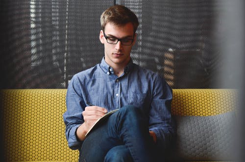 Free Person in Blue Denim Jacket Sitting on Chair While Writing Stock Photo