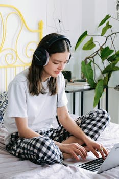 A young woman sits on her bed working remotely on a laptop with headphones on.