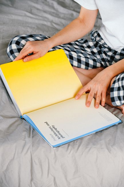 Person reading a book on bed, wearing checked pajamas, relaxing at home.