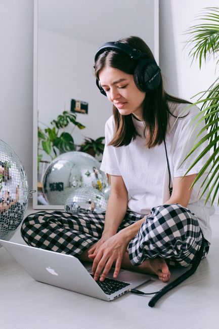 A young woman in a relaxed home setting works on her laptop while listening to music with headphones.