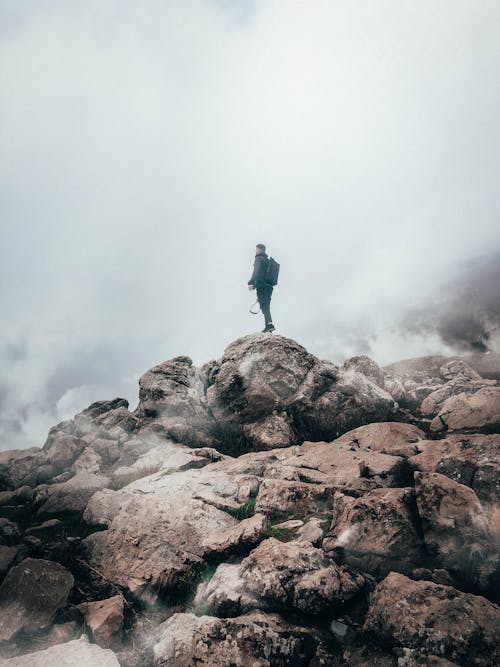 Man in Black Jacket Standing on Brown Rock Formation Under White Clouds