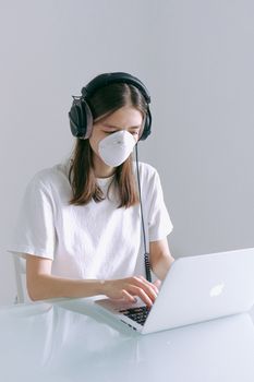 新型冠状病毒肺炎期间，一位戴着口罩和耳机的年轻女子在家里的笔记本电脑前工作