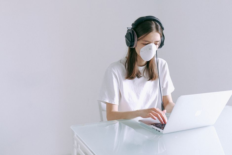Woman wearing a face mask and headphones working on a laptop at home, representing remote work during the pandemic.