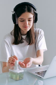 Young woman with headphones using sanitizer while remote working with laptop at home.