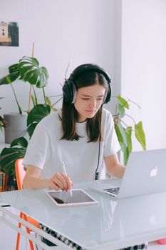 一位年轻女子在家工作，她坐在简约的办公桌前，使用着笔记本电脑、平板电脑和耳机
