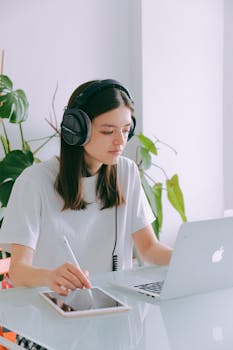 A woman using a laptop and tablet in a cozy home office setting, enjoying music.
