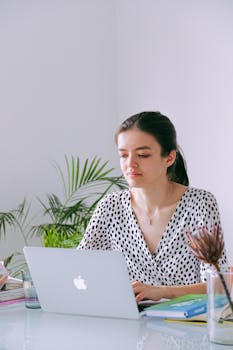 Young woman working on a laptop in a bright home office with plants and natural light.