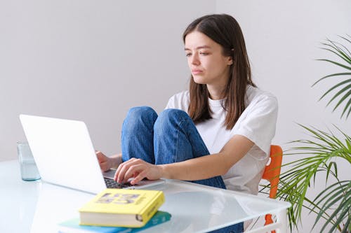 Woman in White Crew Neck T-shirt and Blue Denim Jeans Working on a Laptop