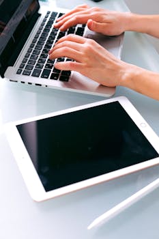 Hands typing on a laptop with a tablet and stylus nearby, showcasing modern technology in a workspace.