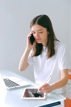A young woman multitasks by using a smartphone while writing notes on a tablet in a bright, modern setting.