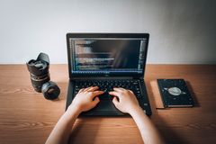 Hands typing on a small laptop with code on screen, a notebook and a camera lens beside it