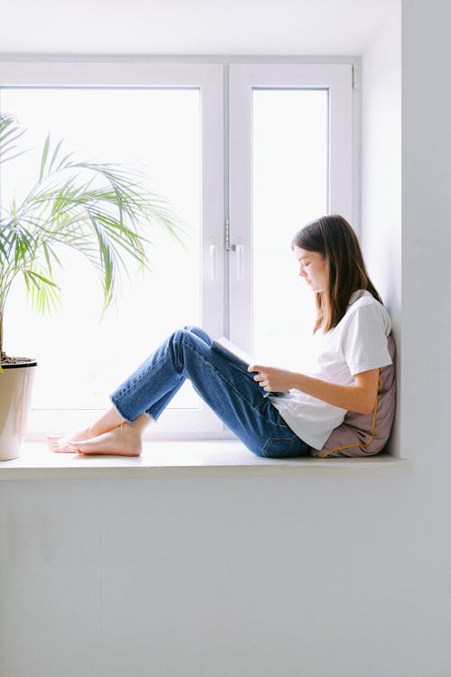 Woman In White Shirt And Blue Denim Jeans Sitting By The Window