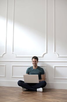 Young man smiling while working remotely on a laptop sitting on the floor at home.