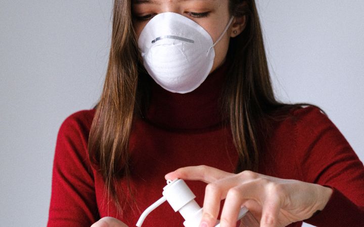 Woman in red sweater applies hand sanitizer wearing a mask indoors for safety and hygiene.