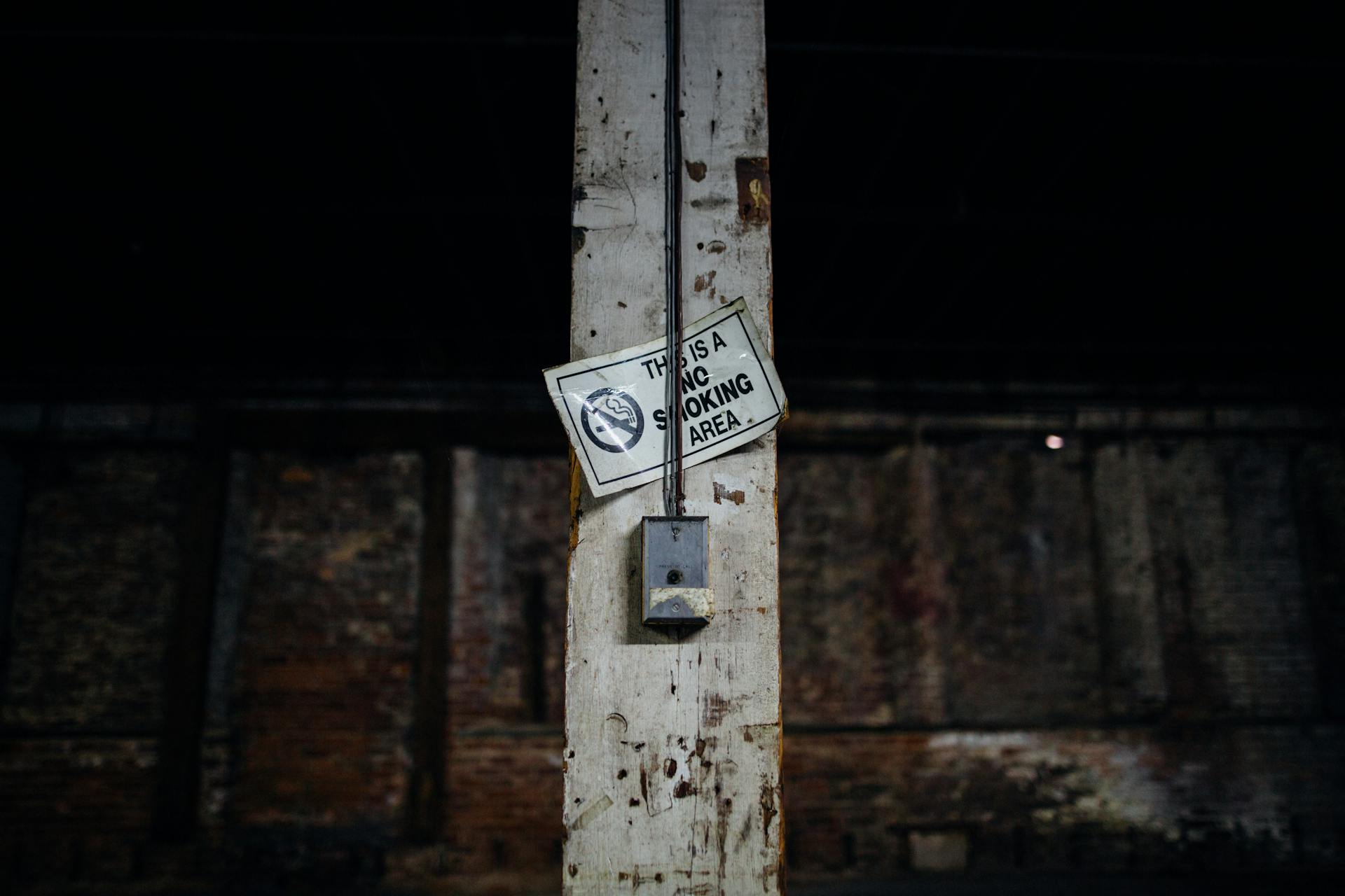 Aged dirty vertical concrete beam with rusty and weathered surface and THIS IS A NO SMOKING AREA title and symbol near rectangular wired device on dark background