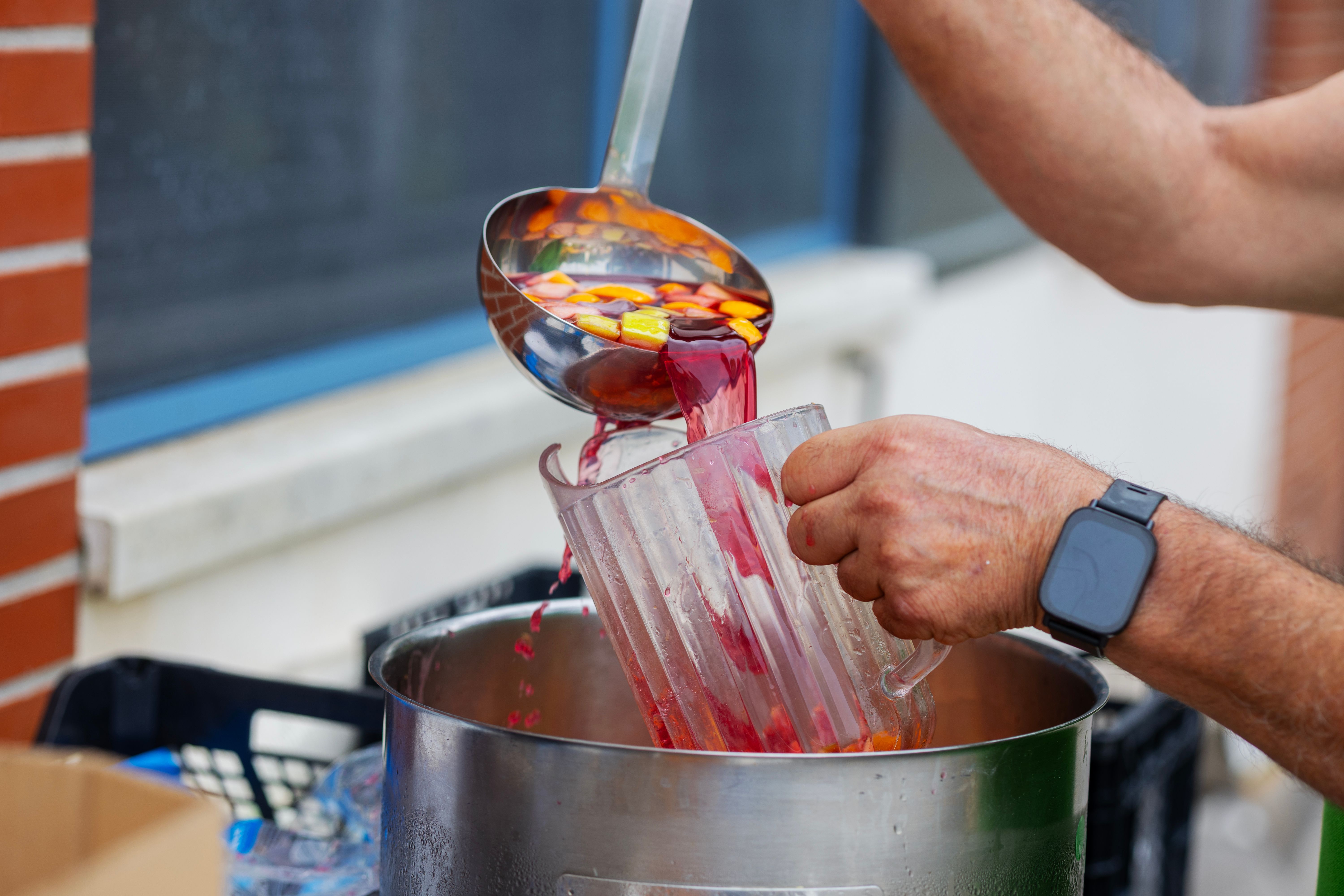 Vibrant fruit punch with fruits being ladled into a transparent pitcher outdoors.