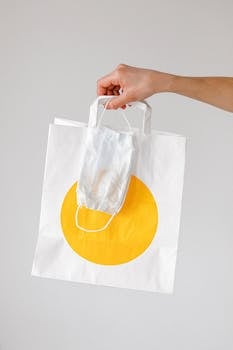 Close-up of a hand holding a paper shopping bag with a face mask, symbolizing pandemic shopping habits.