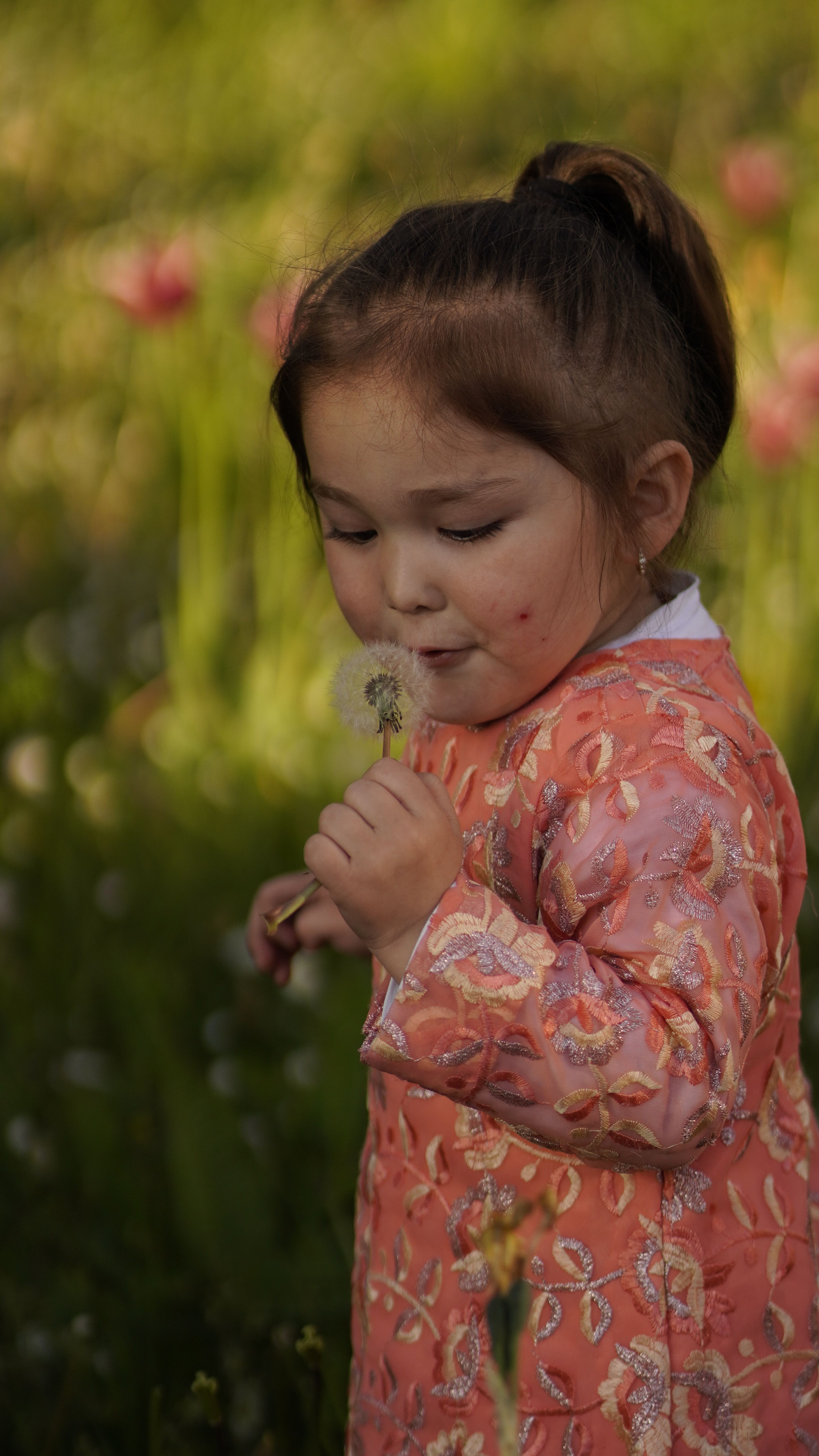 A young girl enjoys a spring day by blowing on a dandelion in a vibrant garden.