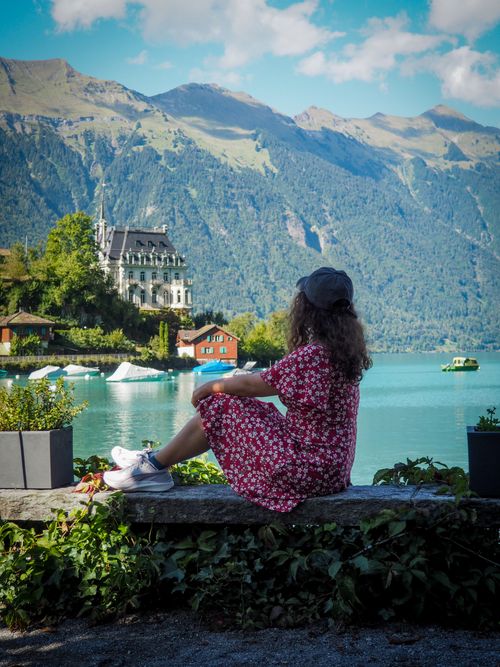 Free Woman in red floral dress sitting by scenic Swiss lake with mountains and historic mansion in view. Stock Photo