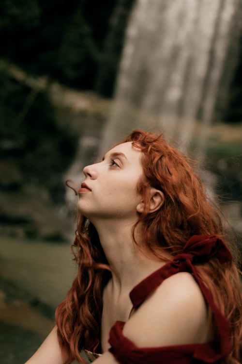 Free A contemplative woman with red hair gazes upwards beside a majestic waterfall. Stock Photo