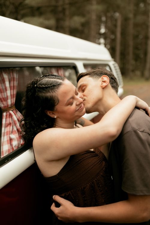 Free A couple sharing a tender moment embracing next to a vintage camper van in a wooded area. Stock Photo