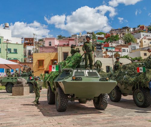 Free Mexican soldiers stand on armored vehicles in a colorful Mexican town square under a clear sky. Stock Photo