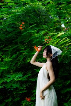 A woman in a white dress poses gracefully amidst vibrant green foliage and delicate orange blossoms.
