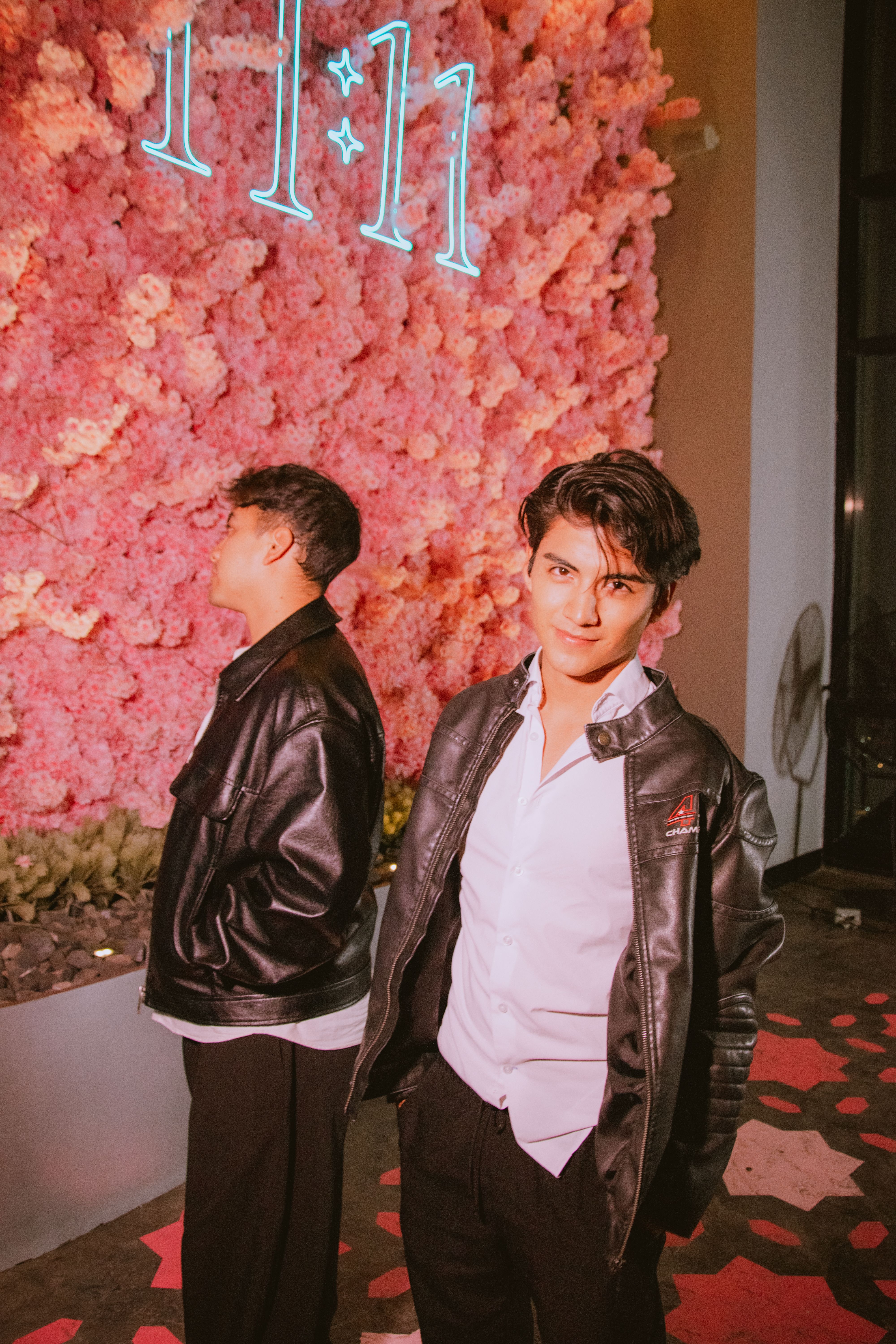Two young men in leather jackets pose indoors with a floral backdrop. Vibrant and stylish setting.