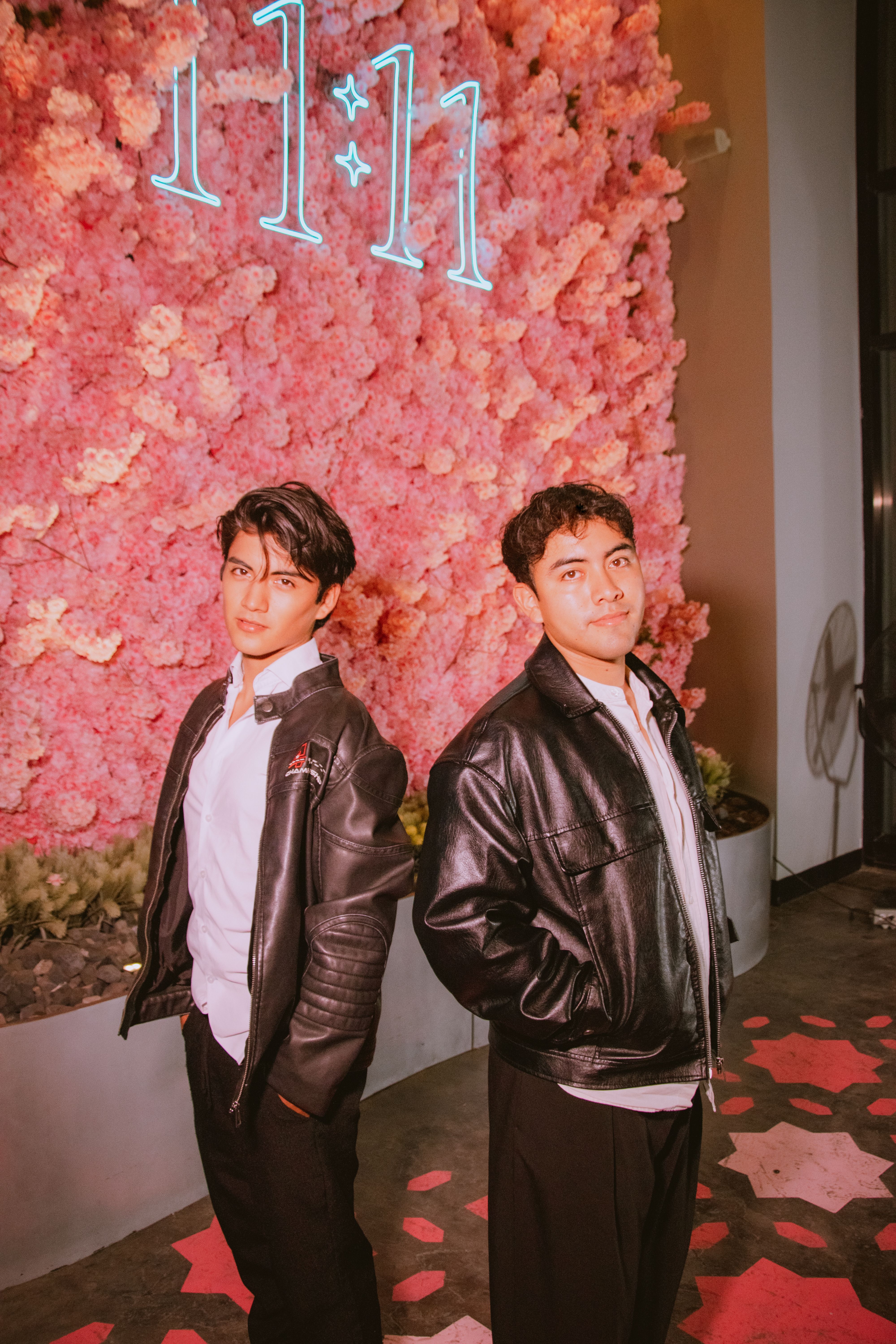 Two young men in leather jackets posing confidently in front of a vibrant pink floral wall.