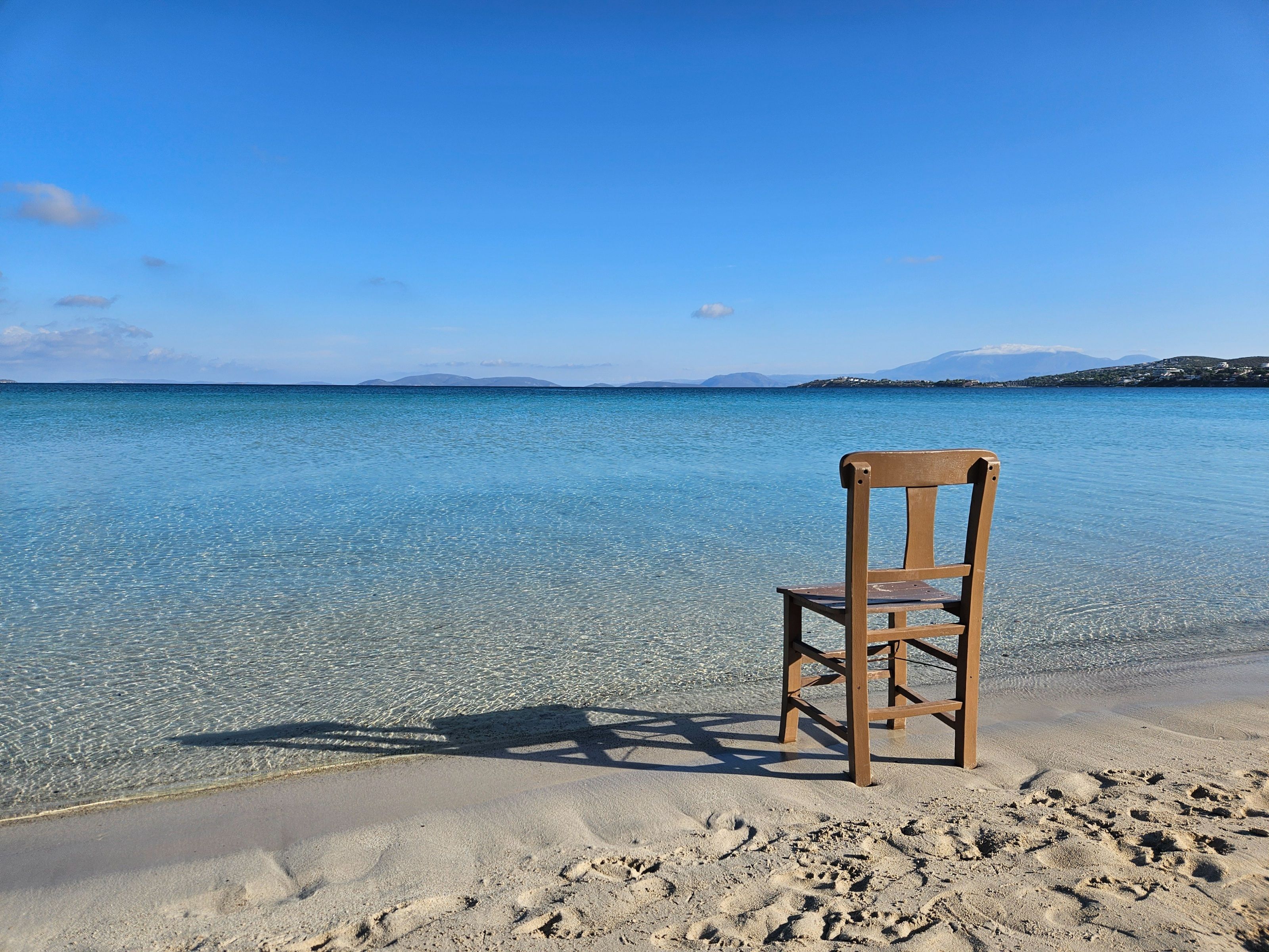 A lone wooden chair sits on a serene beach with clear blue waters and a bright sky.